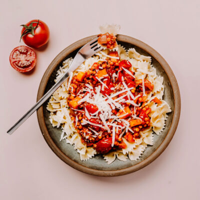 Linsen Bolognese in einer Schüssel mit einer Gabel, im Hintergrund 2 Tomaten und rosafarbener Hintergrund