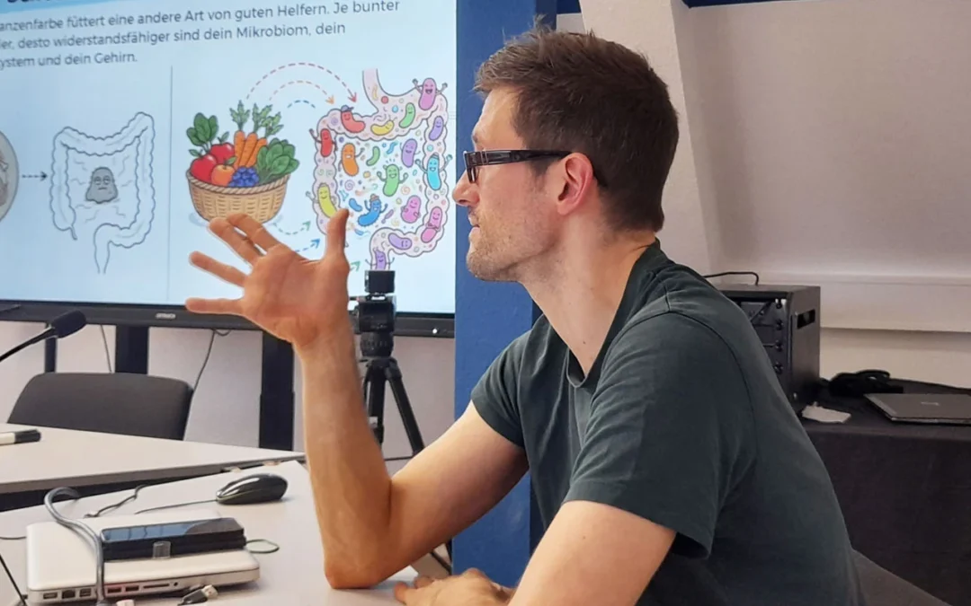 Florian Schaumkessel bei der Lebenshilfe Düsseldorf: Er sitzt am Tisch, schaut geradeaus, im Hintergrund ist seine Präsentation zu sehen.