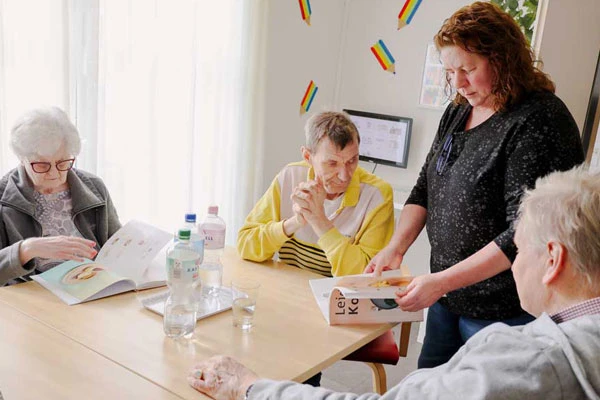 Drei Menschen einer Wohngruppe sitzen am Tisch, eine Betreuerin zeigt ihnen ein Kochbuch, durch das sie gerade blättert